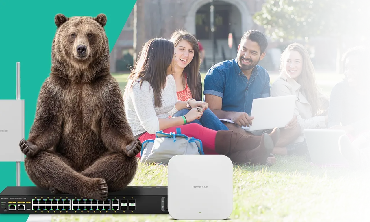 A grizzly bear sitting on a NETGEAR switch beside NETGEAR access points, with students working on laptops on a campus lawn